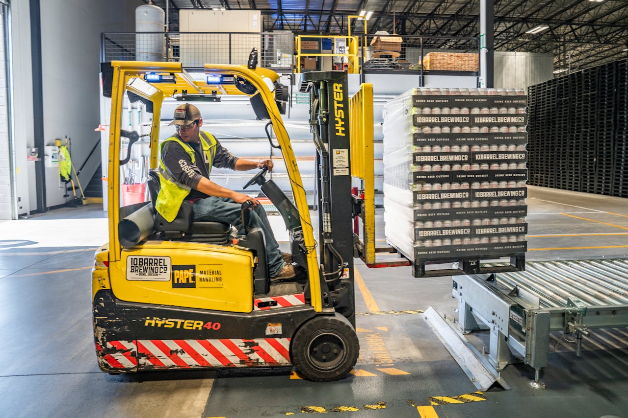 creative-03 A warehouse worker maneuvers a forklift to transport crates for brewing company storage.
